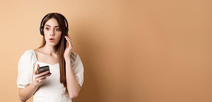 Wow awesome tunes. Excited young girl amazed with new song, listening music in headphones and look impressed, holding cell phone, beige background.