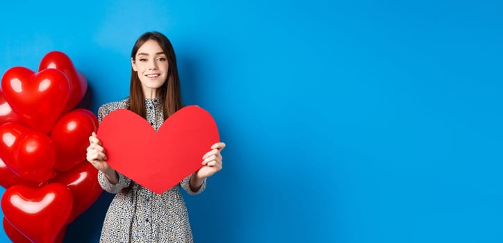 Valentines day. Lovely young woman in dress celebrating lovers holiday, showing valentine card and smiling, standing near red heart balloons on blue background.