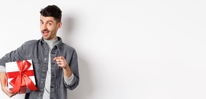 Portrait of romantic guy buying gift for Valentines day and pointing at you, inviting to special deal shopping, standing with present on white background.