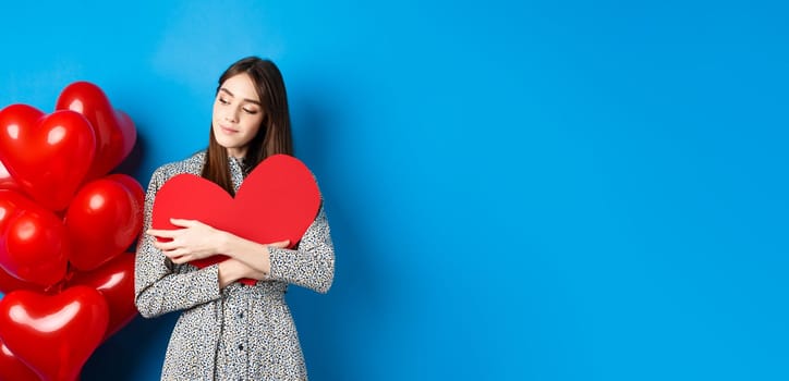 Valentines day. Romantic pretty woman in dress hugging big red heart cutout and looking dreamy, thinking of love, standing on blue background.