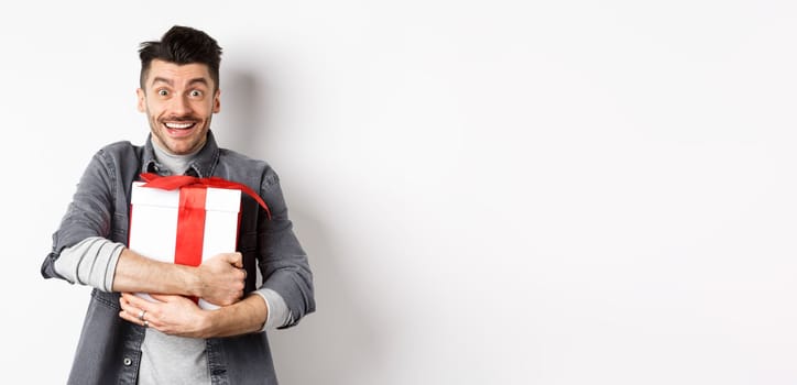 Happy caucasian man holding surprise gift, hugging present and smiling thankful, looking at camera with love and appreciation, celebrate valentines holiday, white background.