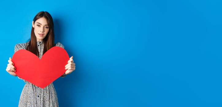 Valentines day and relationship concept. Tender young woman in dress stretch out hand and giving big red heart to you, making confession, standing romantic on blue background.