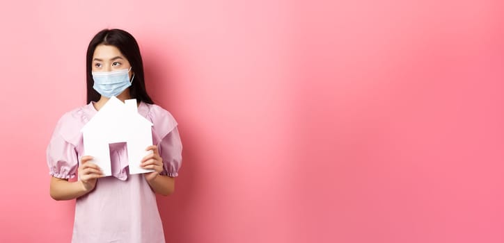 Real estate and pandemic concept. Young asian woman in medical mask dreaming of home, showing paper house cutout and look aside, standing against pink background.