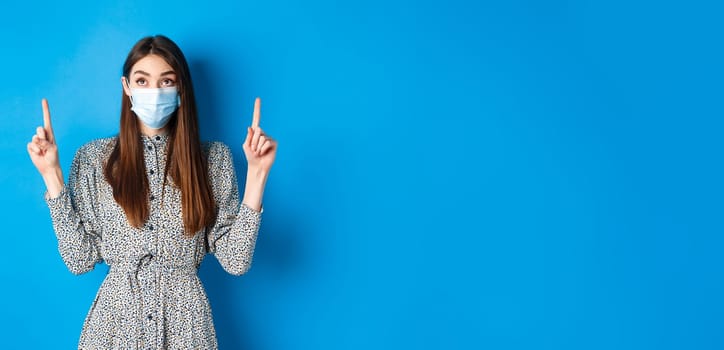 People, covid and quarantine concept. Intrigued young woman looking and pointing up at advertisement, standing in dress and face mask from coronavirus, blue background.