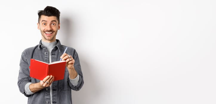 Creative young man smiling excited while taking notes, writing down ideas in planner, holding red journal and looking happy, standing against white background.