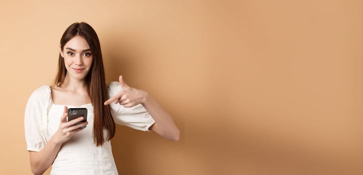 Attractive woman smiling and pointing at smartphone, showing online offer on phone, standing on beige background.