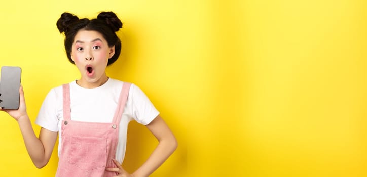 Online shopping concept. Excited stylish asian girl say wow, showing empty cell phone screen and look amazed, yellow background.