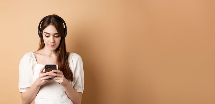 Woman chatting on mobile phone and listening music in wireless earphones, standing on beige background.