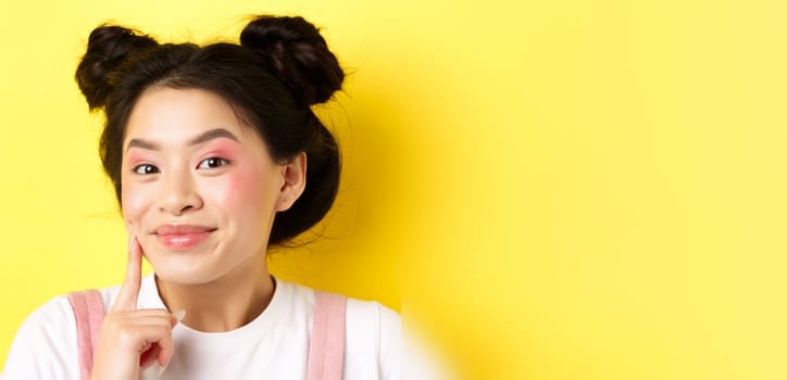 Beauty. Stylish asian girl with bright glamour makeup, touching soft and shiny facial skin, smiling happy at camera, standing on yellow background.