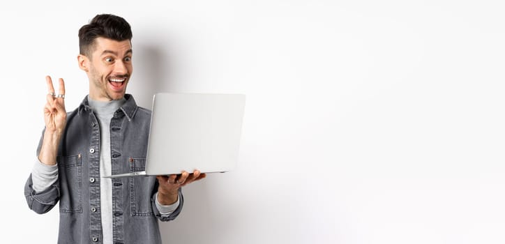 Funny young man video chat on laptop, showing v-sign and smiling at computer camera, standing against white background.