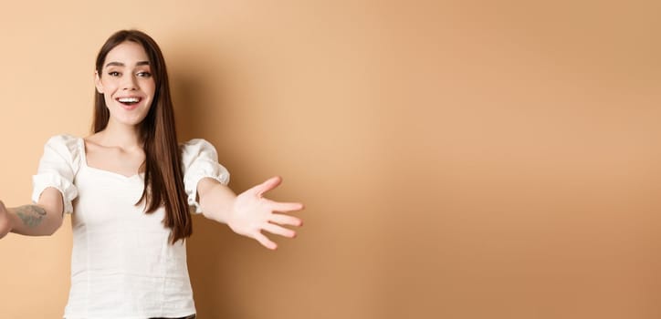 Portrait of friendly candid girl reaching for hug, welcome friend and smiling, spread hands sideways in greeting gesture, receiving something, beige background.
