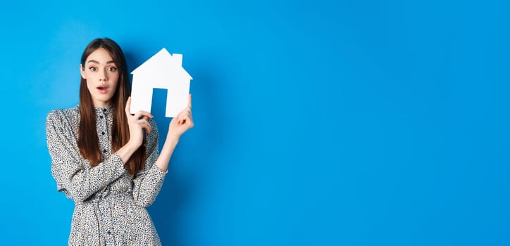 Real estate, realtors and insurance concept. Excited woman gasping and holding paper house cutout, see good deal, standing on blue background.