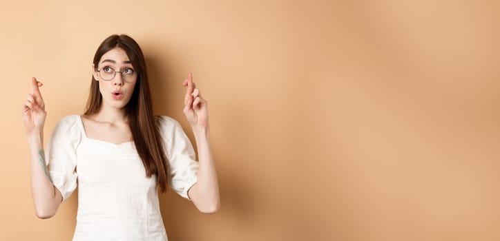 Excited girl hope dream come true, making wish with fingers crossed, looking aside and praying, standing on beige background.