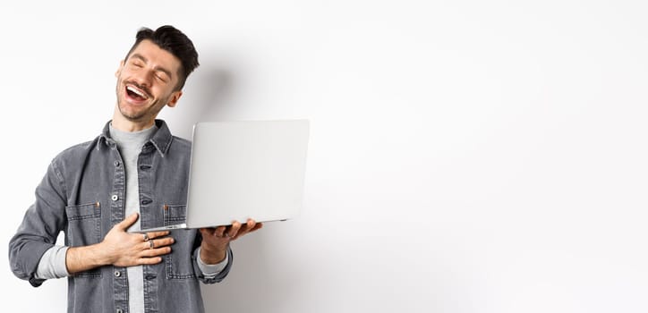 Image of happy guy laughing at funny video on laptop, touching belly and smiling satisfied wth good laugh, standing against white background.