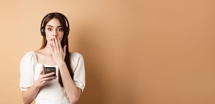 Surprised young woman gasping and covering mouth with hand, using wireless headphones to listen podcast or music, holding mobile phone, beige background.