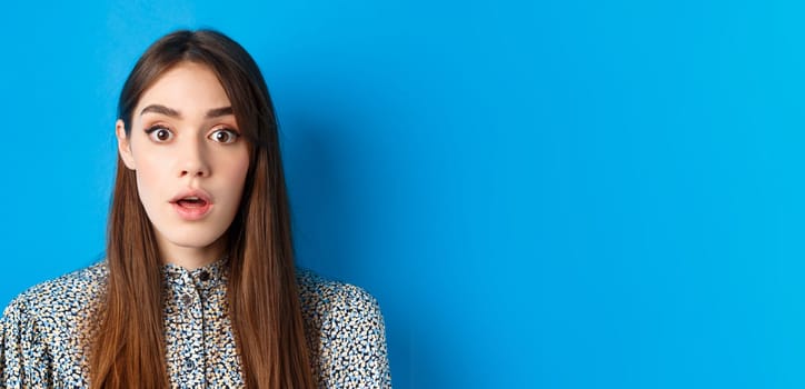 Close-up of excited pretty woman with long natural hair and makeup, drop jaw and gasping surprised, checking out awesome promo, hear amazing news, blue background.