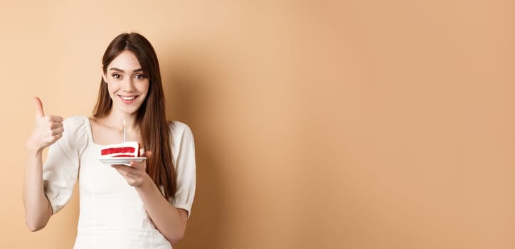 Beautiful birthday girl making wish on cake, showing thumb up and smiling happy, enjoying b-day party, standing on beige background.