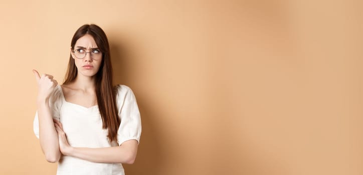 Suspicious woman in glasses pointing and looking aside, frowning with doubtful face, standing on beige background.