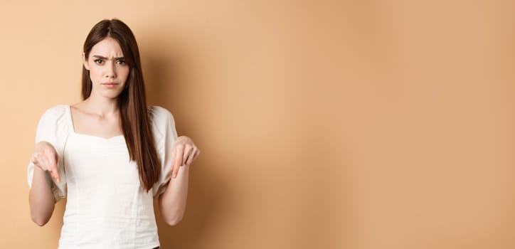 Skeptical young woman frowning, pointing hands down at something bad or disappointing, look upset, standing on beige background.