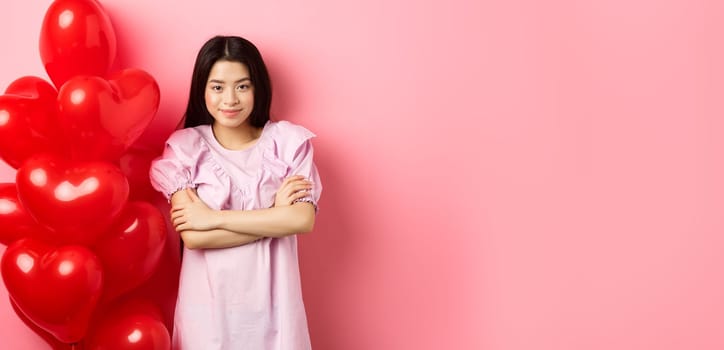 Confident teenage girl cross arms on chest and smile, celebrating valentines day in cute dress with red heart balloons, standing on pink background.