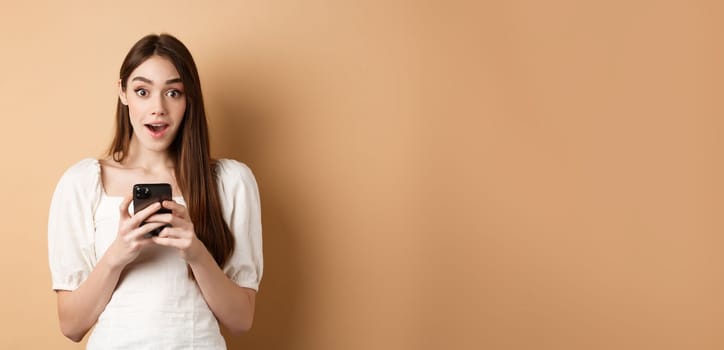 Exciting news on phone. Amazed young woman say wow after reading online announcement on smartphone, standing on beige background.