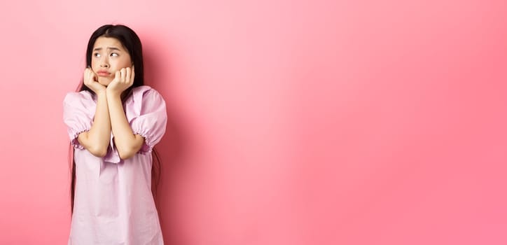 Sad and lonely asian girl look left and frowning, standing distressed against pink background.