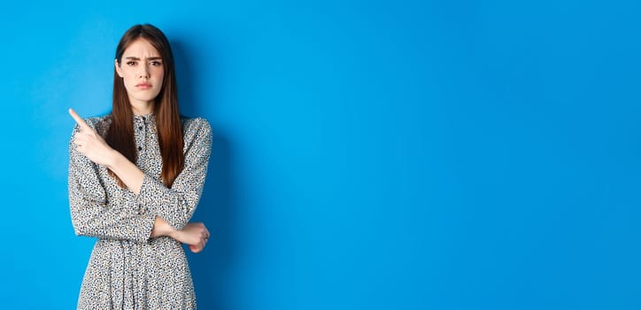 Disappointed frowning girl pointing left and looking displeased, condemn something bad, complaining at banner, standing against blue background in dress.