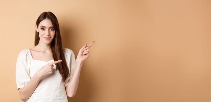 Beautiful young woman in dress pointing fingers right, showing logo and looking tender at camera, beige background.