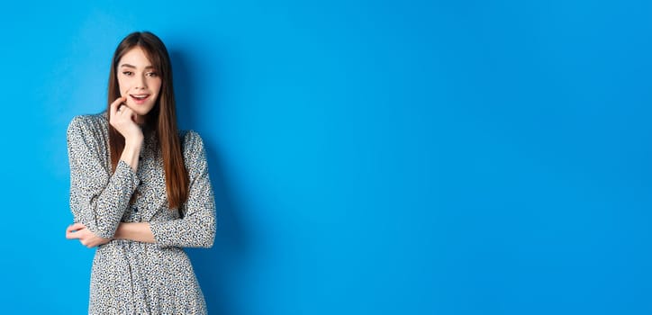 Coquettish girl in trendy long dress looking excited at camera, standing on blue background.