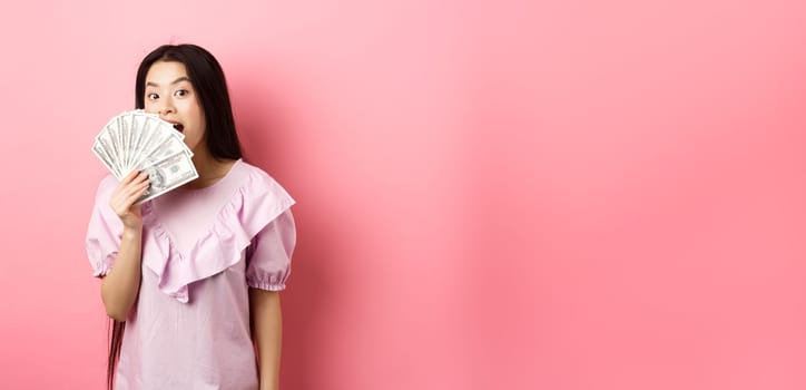 Cute asian woman showing dollar bills and smiling amazed. Rich woman waiving with money, standing against pink background.