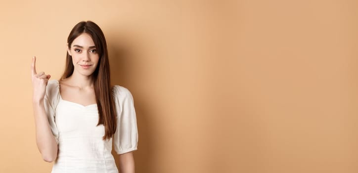 Attractive young woman show finger number one, smiling and looking confident, standing on beige background.