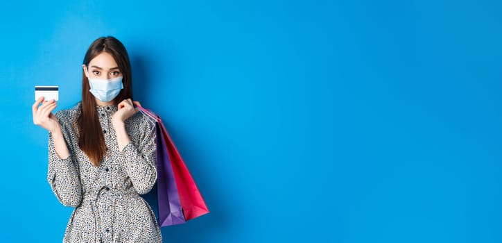 Covid-19, pandemic and lifestyle concept. Stylish woman wearing medical mask on shopping as preventive measure from coronavirus, showing plastic credit card and holding bags, blue background.