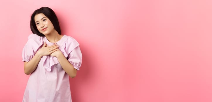 Dreamy asian girl smiling and holding hands on chest, looking left at something romantic and cute, standing on pink background.