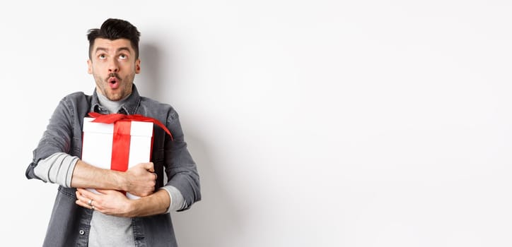 Surprised funny guy hugging big holiday gift and looking up, say wow amazed, checking out Valentines day special offer, standing on white background.