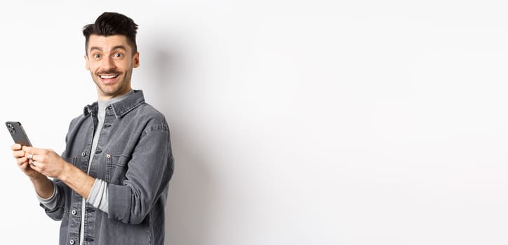 Handsome young man using smartphone and looking at camera. Caucasian guy chatting on phone with happy face, standing on white background.