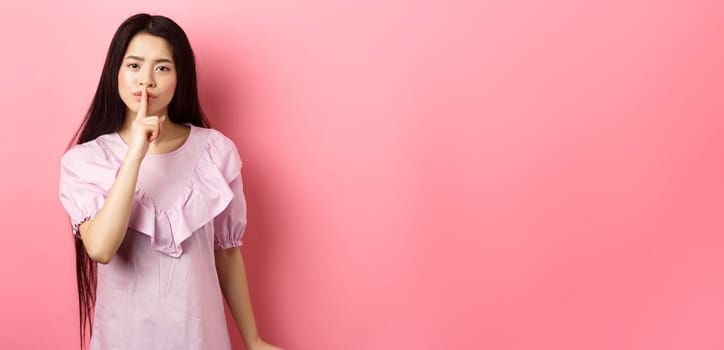 Cute asian girl tell to be quiet, scolding loud person, showing shush sign with finger pressed to lips, standing in dress on pink background.