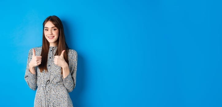 Cheerful smiling woman in dress, showing thumb up and look satisfied, approve and like good thing, recommending product, standing on blue background.