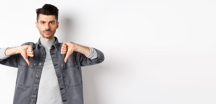 Very bad. Serious man showing thumbs down and look at camera with pokerface, telling no, disapprove and dislike product, standing dissatisfied on white background.