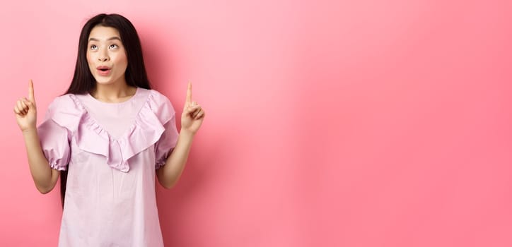 Excited teen asian woman pointing fingers up, looking dreamy at empty space, standing in dress against pink background.