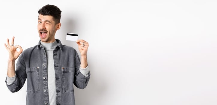 Very good. Smiling guy with plastic credit card showing okay sign, smiling and winking satisfied, recommend bank, standing on white background.