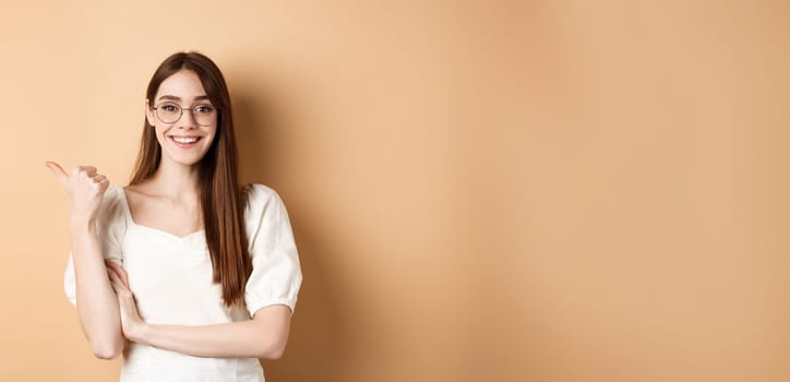 Cheerful woman in eyewear pointing finger left at logo, smiling happy, standing on beige background.