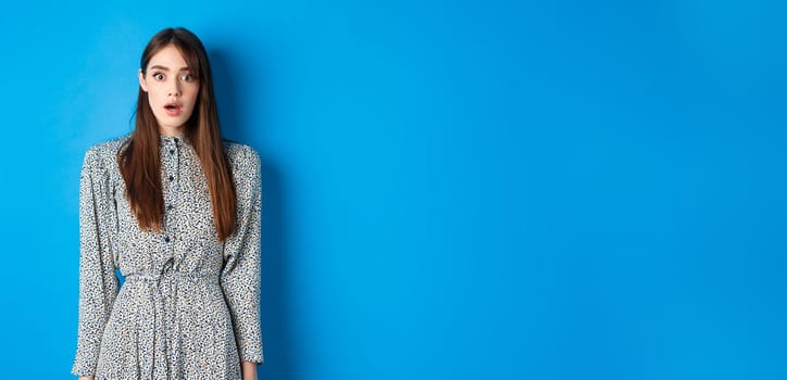 Shocked cute girl in dress looking at camera with opened mouth, gasping startled, see something shocking, standing on blue background.