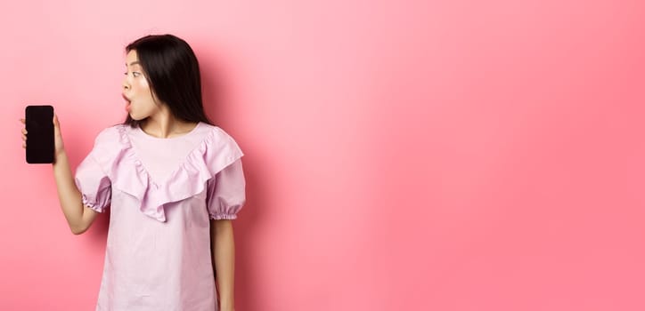 Wow look here. Excited asian girl look at smartphone screen with amazed face, checking out online offer, standing against pink background.