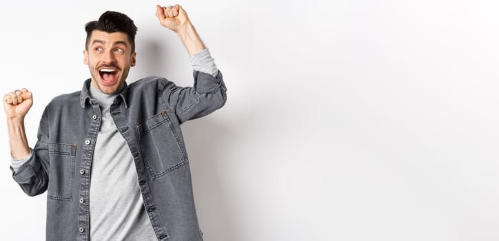 Excited young man cheering and looking aside, winning prize lottery, making good deal and celebrating, achieve goal or success, do victory dancy and shout yes, white background.