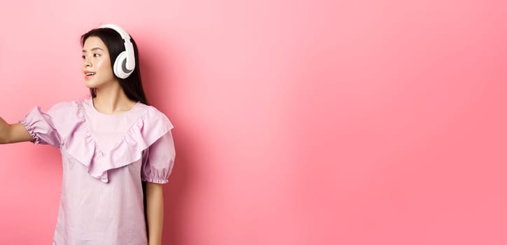 Beautiful asian girl blogger taking selfie in wireless headphones, make photo for social media on smartphone, standing in dress against pink background.