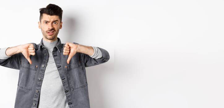 Disappointed guy showing thumbs down and frowning, looking skeptical at something bad, dislike and disapprove awful thing, standing on white background.