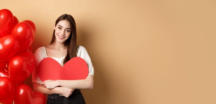 Valentines day and love concept. Lovely girl hugging big romantic heart cutout and smiling, standing near red balloons on beige background.