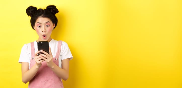 Asian teen girl look excited at smartphone screen, reading news on phone, standing on yellow background.