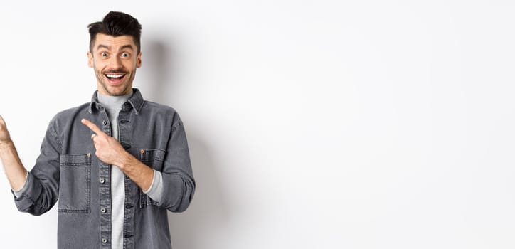 Excited guy with moustache pointing fingers left, showing advertisement and smiling happy, standing on white background.