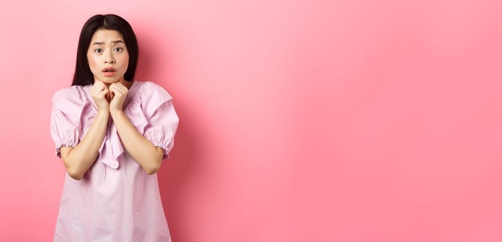 Scared timid girl looking worried at camera, standing concerned on pink background.